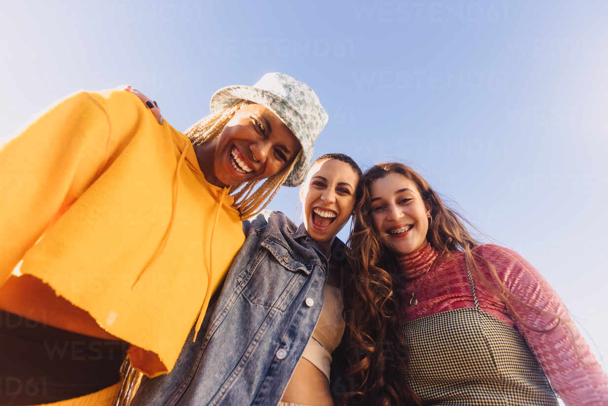Low angle view of three friends smiling at the camera cheerfully. Female youngsters having fun while standing outdoors in the city. Group of generation z friends making happy memories together.